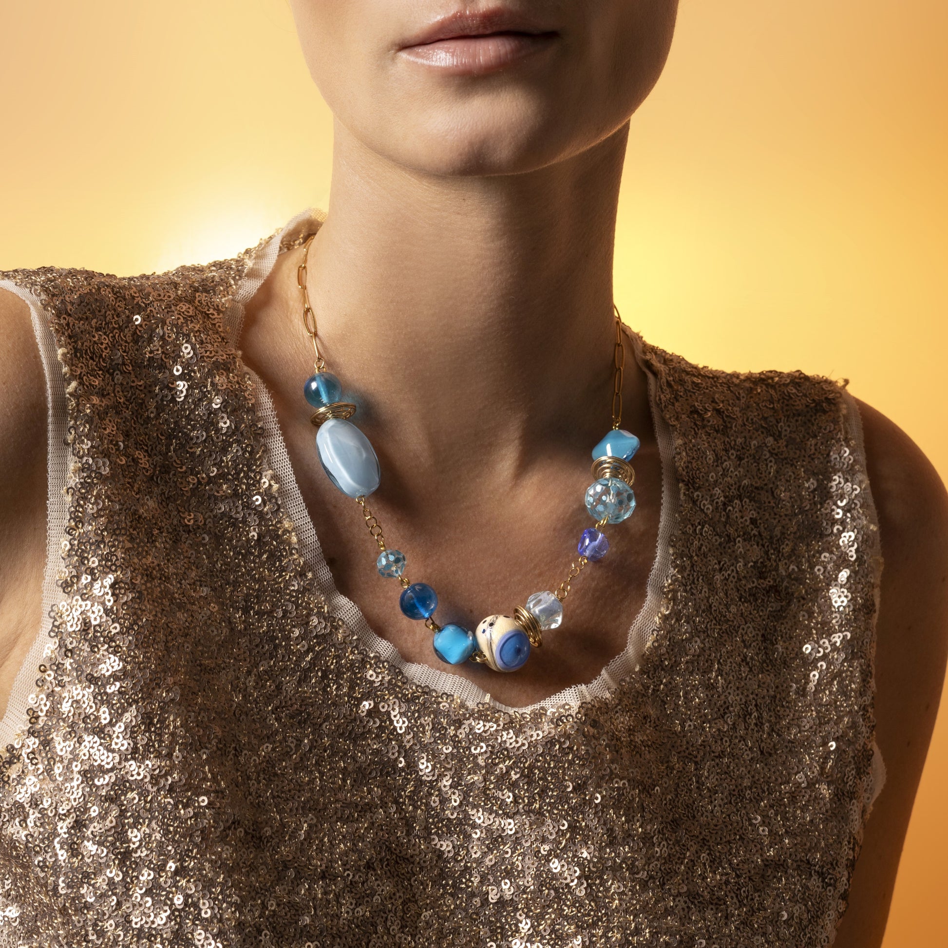 Glow Murano glass necklace with 18KT Gold Finish over brass, adjustable 18 inches plus extension, handcrafted in Venice, Italy. worn by a person in a sparkly top against an orange background